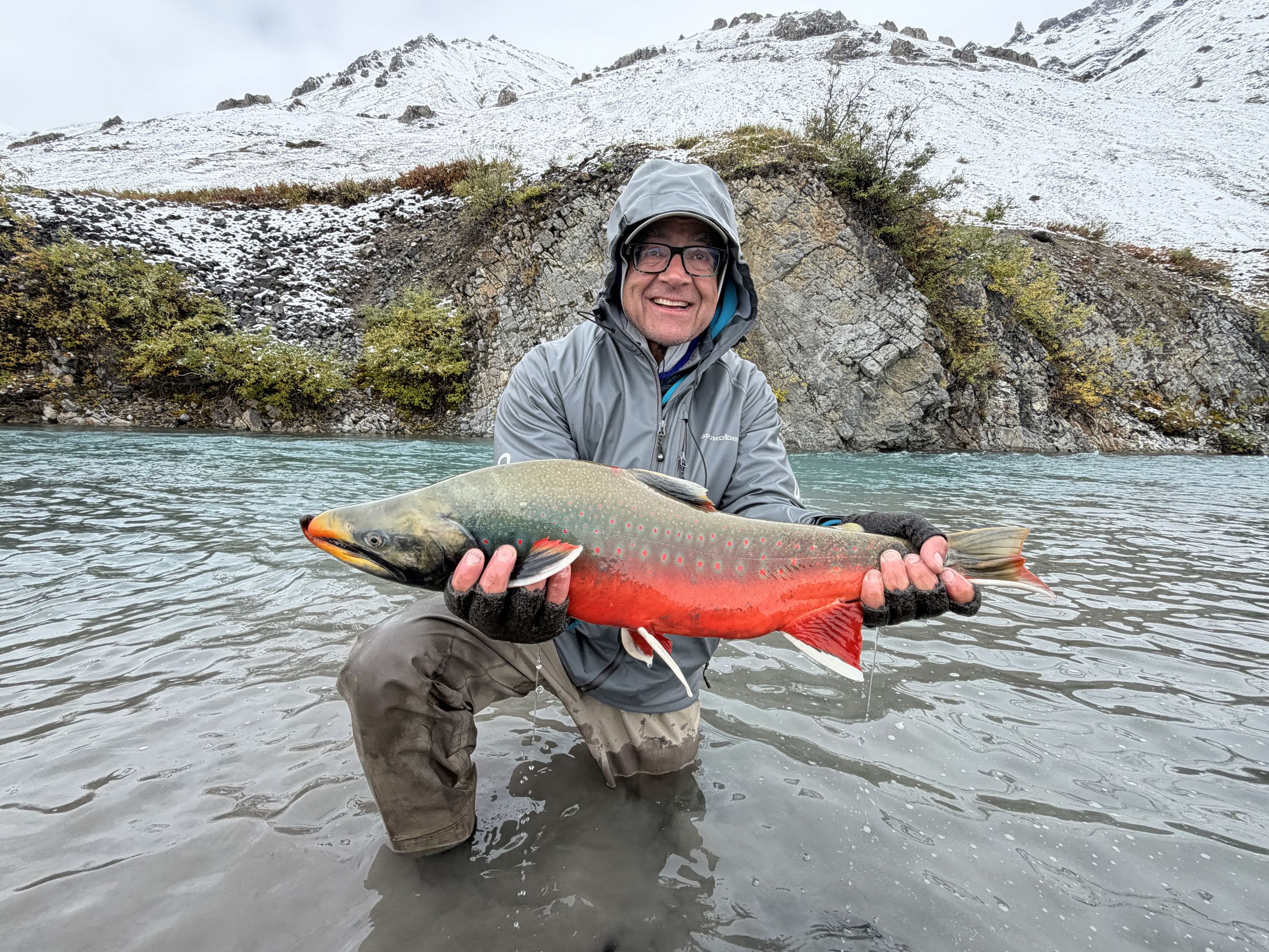A brilliantly colored, spawning-phase Dolly Varden held above the water on the Ivishak River, with fresh snow on the surrounding mountains.