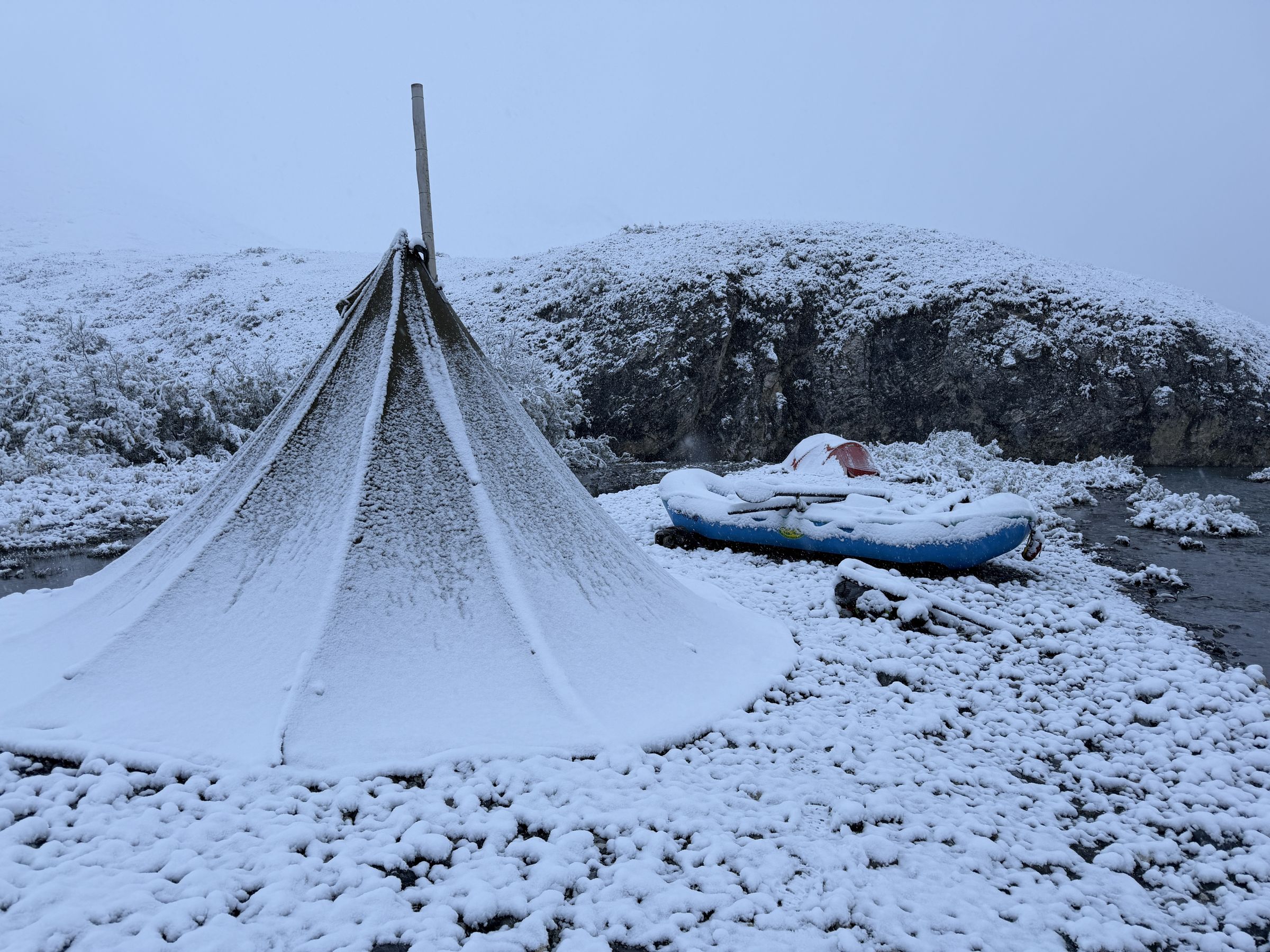 A snow-covered tipi and raft at a Remote Waters wilderness camp on the Ivishak River during an early-season weather event.