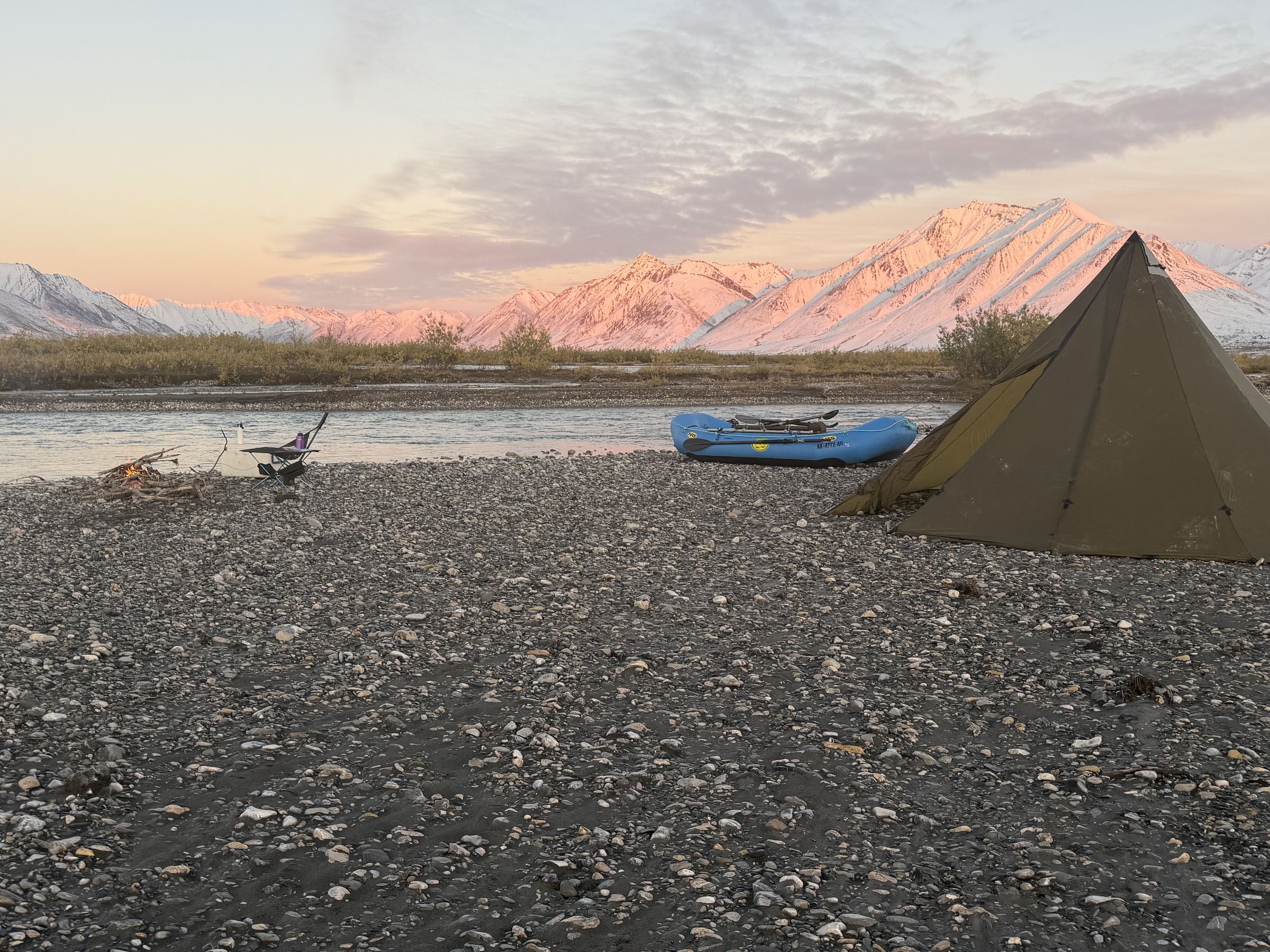 A tipi and raft on a gravel bar at sunset on the Ivishak River, with snow-covered Brooks Range peaks behind camp.
