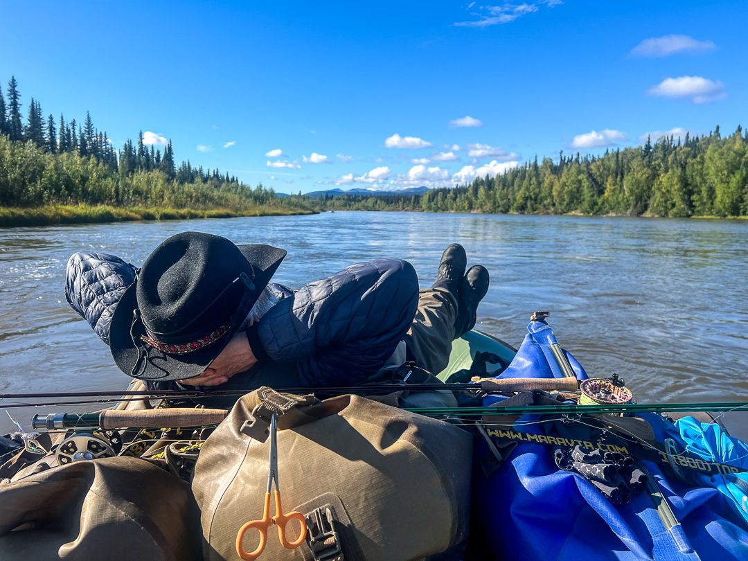 An angler reclines on a loaded raft while floating the Kobuk River, fishing equipment in the foreground and river and forest extending into the distance.