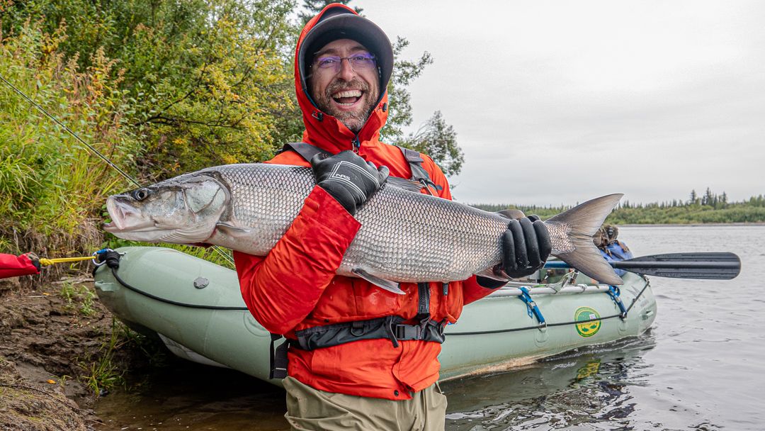 A smiling angler in a red jacket holds a large sheefish beside a raft on the Kobuk River.
