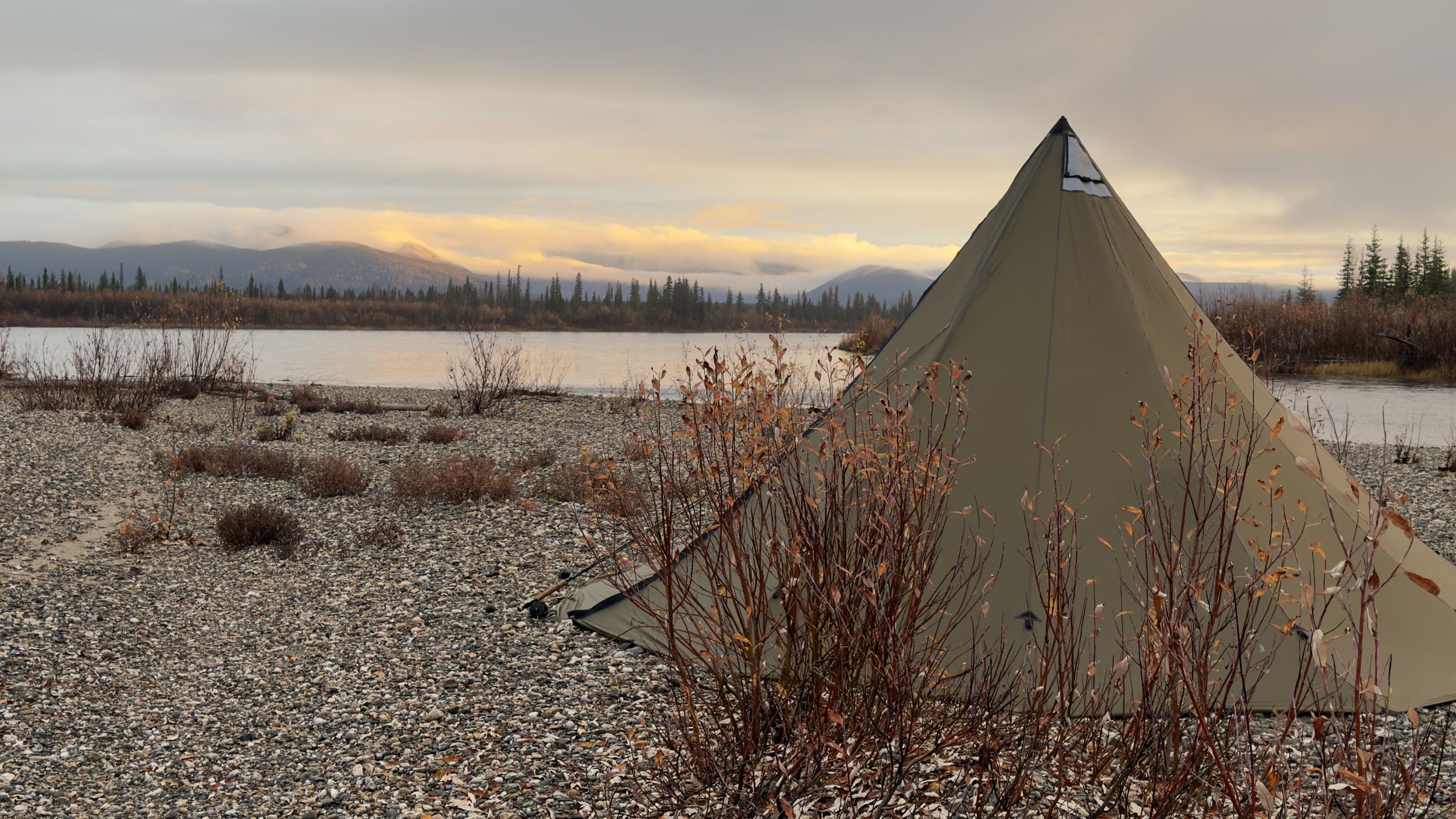 A tipi-style tent on a gravel riverbank at evening, with the river, autumn brush, and mountains lit by sunset.