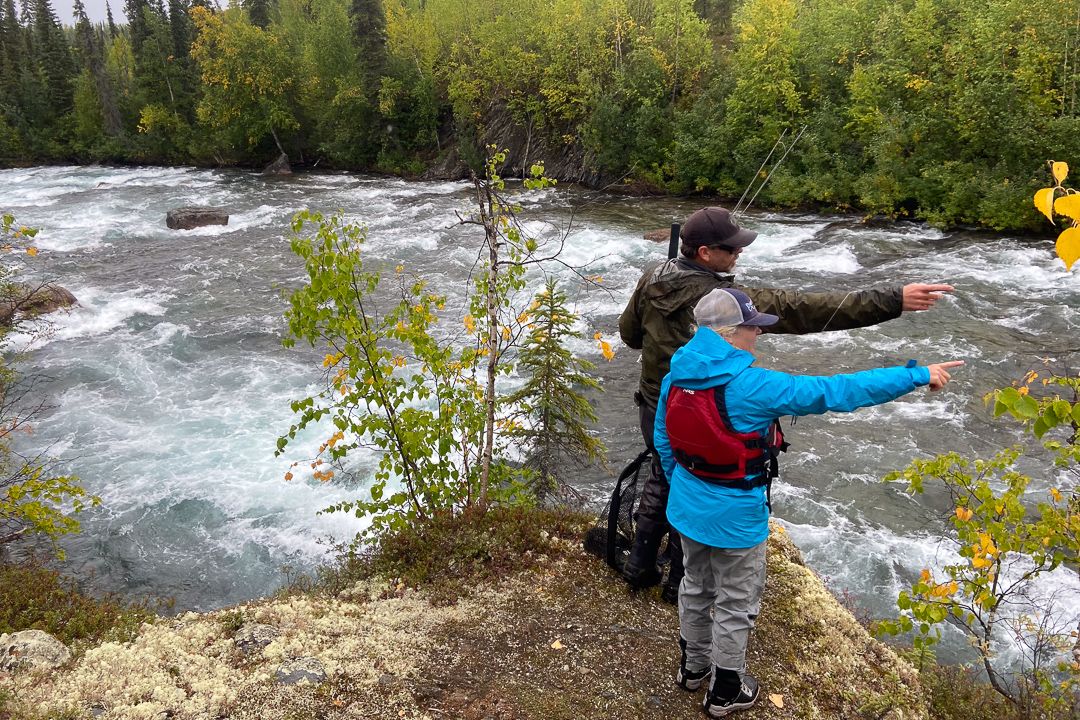 Two people scout an upper rapid on the Kobuk River, one pointing out a line through the water.
