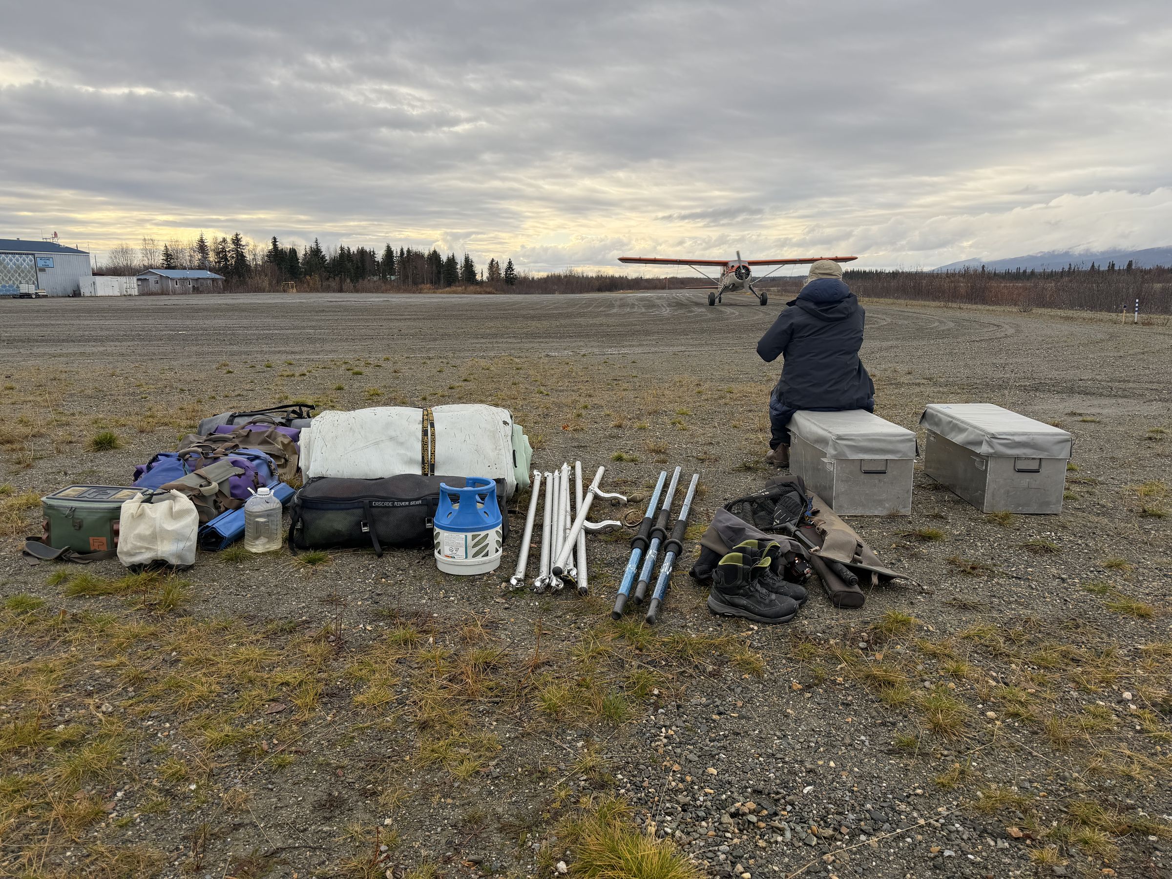 Dry bags, supply boxes, a propane canister, and other gear staged on a gravel airstrip, with a bush plane taxiing in the background.